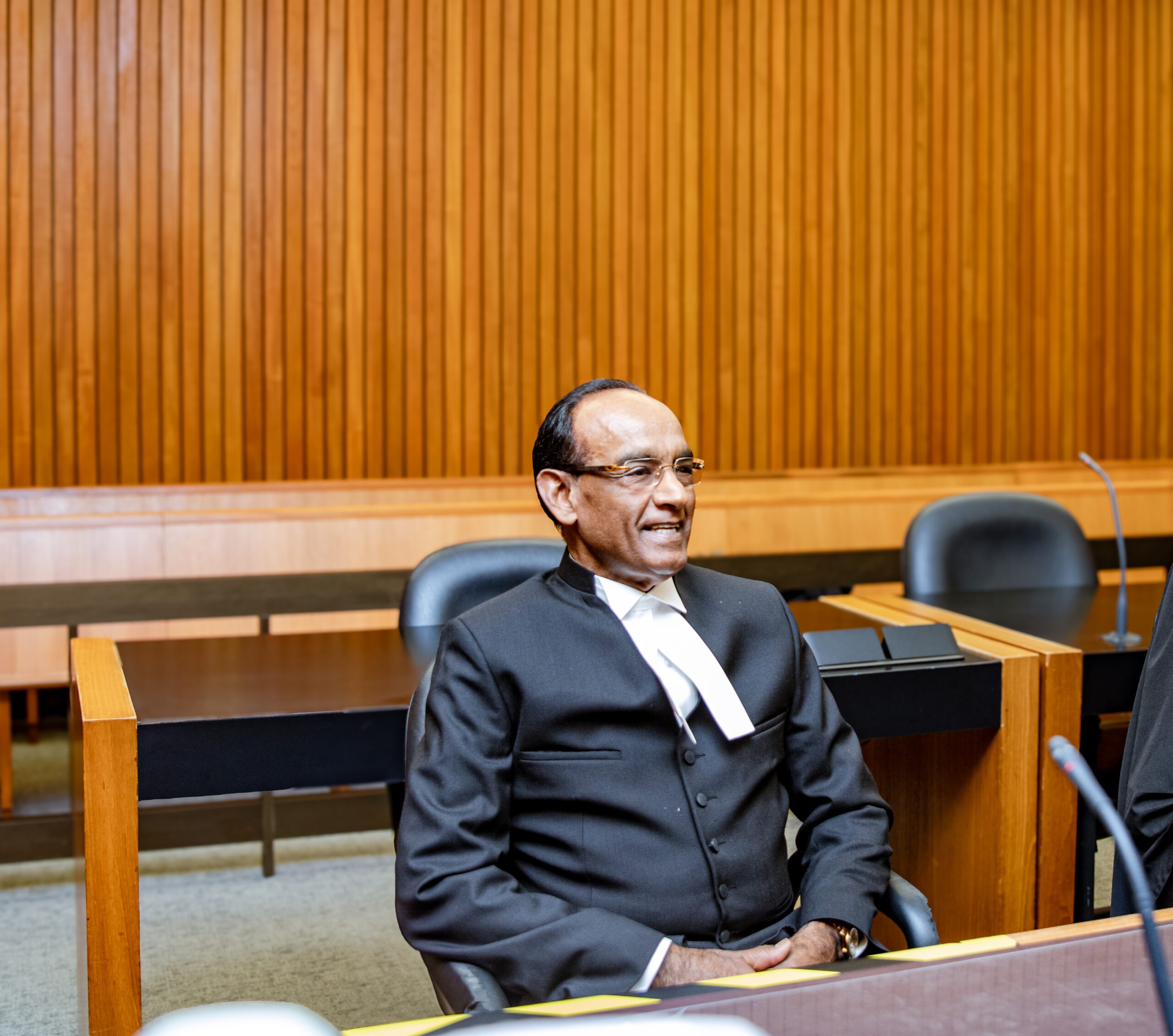 A smiling judge in formal attire sits in a courtroom, with wooden paneling in the background, emphasizing the judicial context.