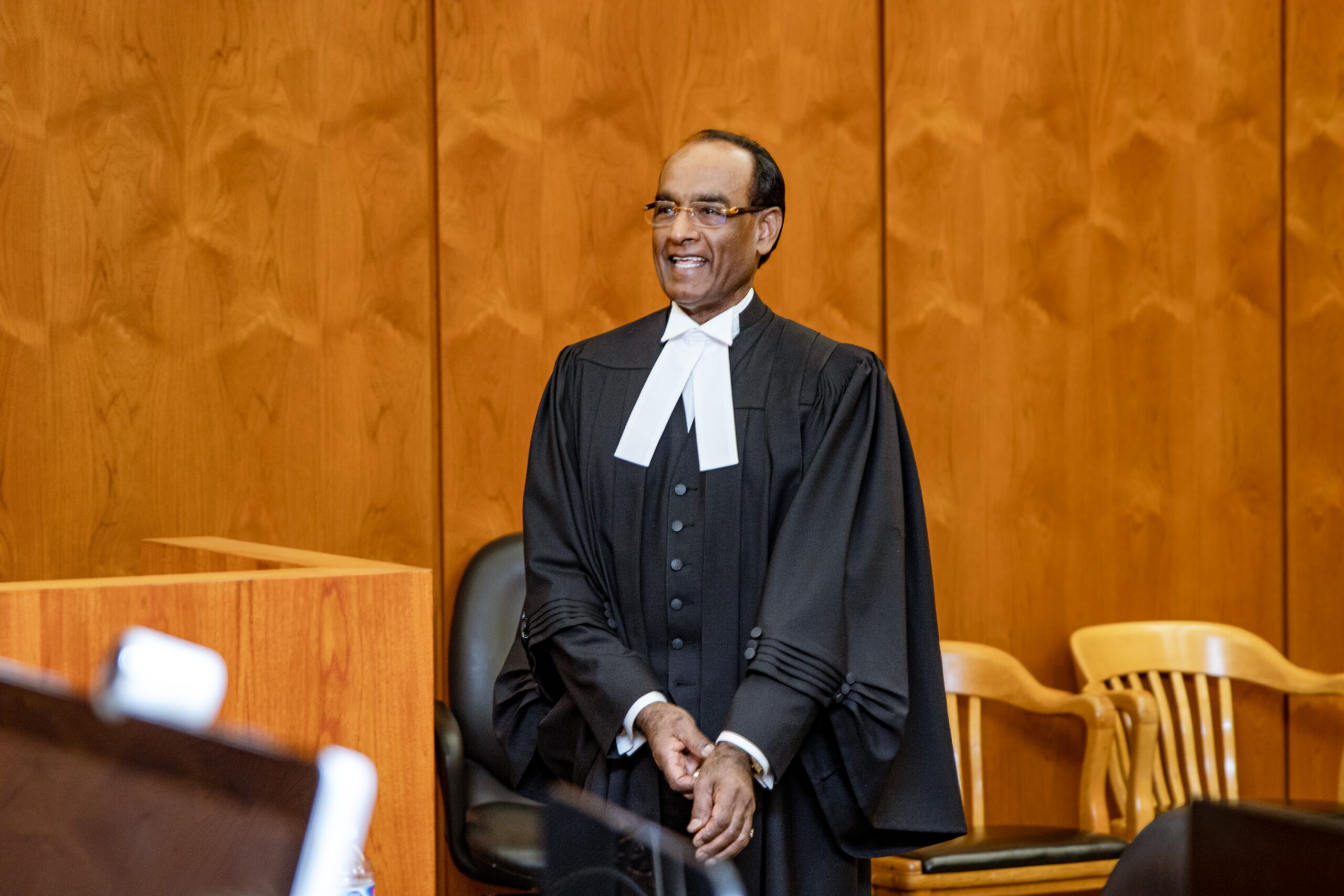 A smiling judge stands in a courtroom, wearing a black robe and white collar, against a wooden panel backdrop, conveying a sense of authority and professionalism.